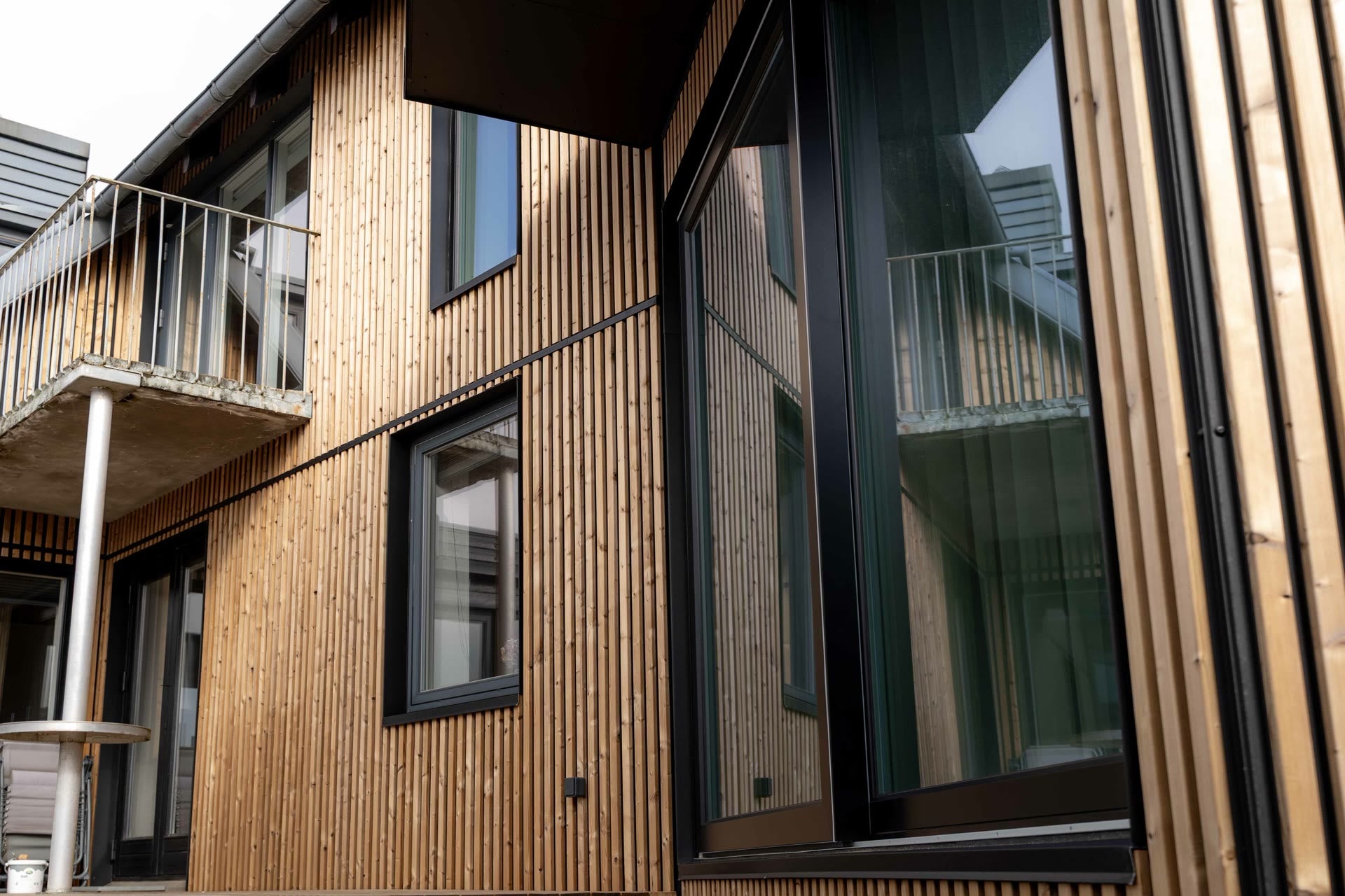 Angled view of a house facade clad in vertical oiled Aluwood slats with a balcony and black window frames